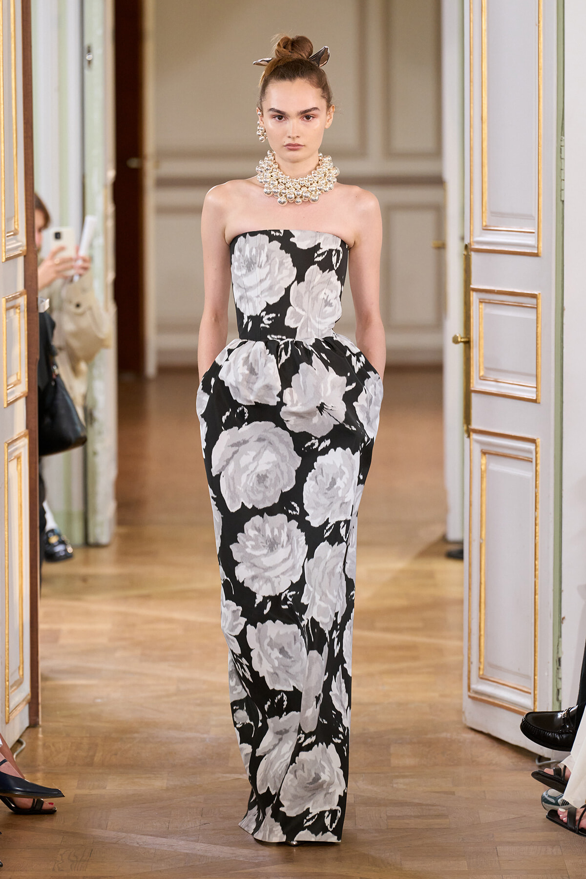 Model wearing a strapless black-and-white floral gown with a voluminous skirt and a chunky pearl necklace, walking in an ornate doorway hallway.