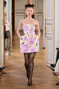 Model walking the runway in a strapless pastel floral mini dress with purple flowers, paired with black lace tights and black patent heels.