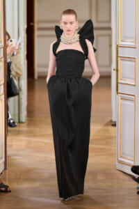 Model in a black strapless gown with ruching, a voluminous bow-shoulder detail, and a pouched skirt, wearing a chunky pearl necklace in a gilded room doorway.