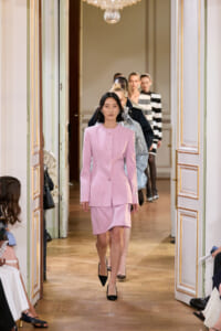 Model walks in pastel pink tailored suit with blazer and skirt, down a chandelier-lit, ornate hallway runway.
