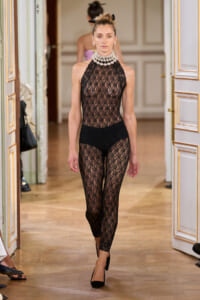 Model walking a runway in a black lace high-neck bodysuit with matching lace leggings and large pearl choker in an ornate doorway setting