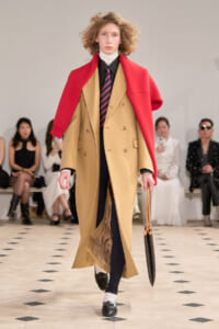 Model walks runway in a tan double-breasted coat with a bright red cape draped over shoulders, paired with a striped tie and black shirt beneath a white turtleneck, holding a patterned umbrella bag.