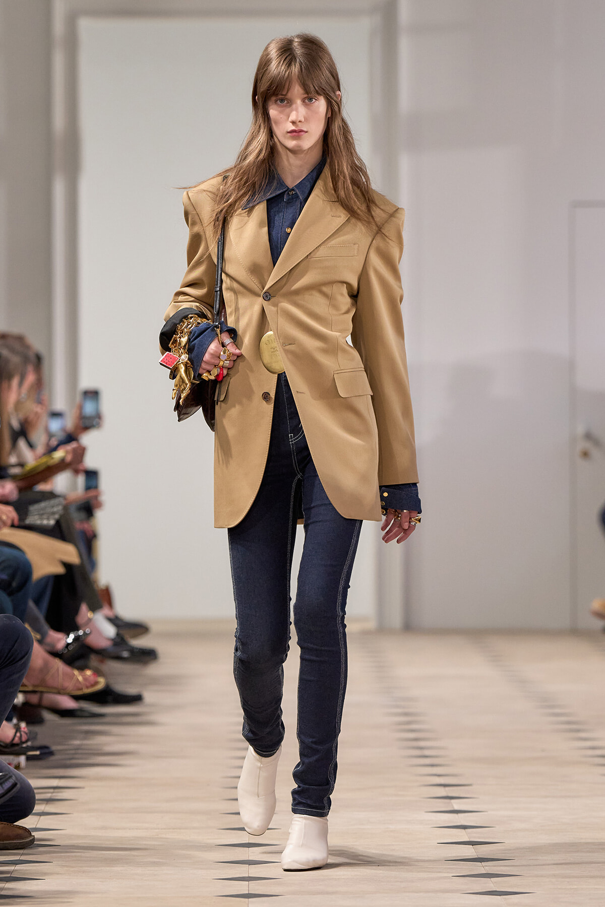 Model walks the runway in a camel blazer over a blue denim shirt and dark jeans, with white ankle boots and a jeweled handbag.Paraphrase option 2 not needed.