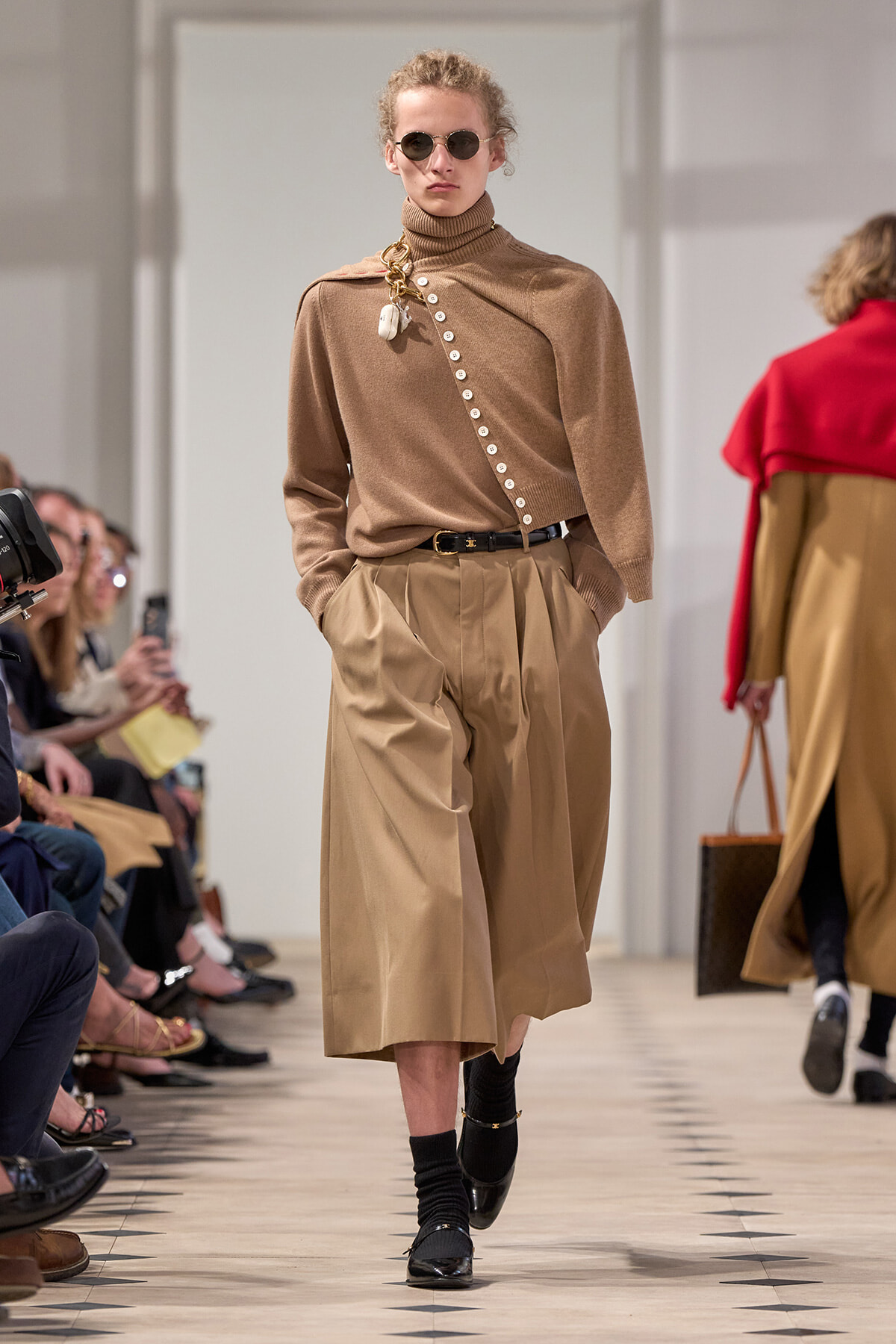 Model walking the runway in a camel turtleneck with a diagonal row of white buttons and wide beige culottes, accessorized with a belt and sunglasses.