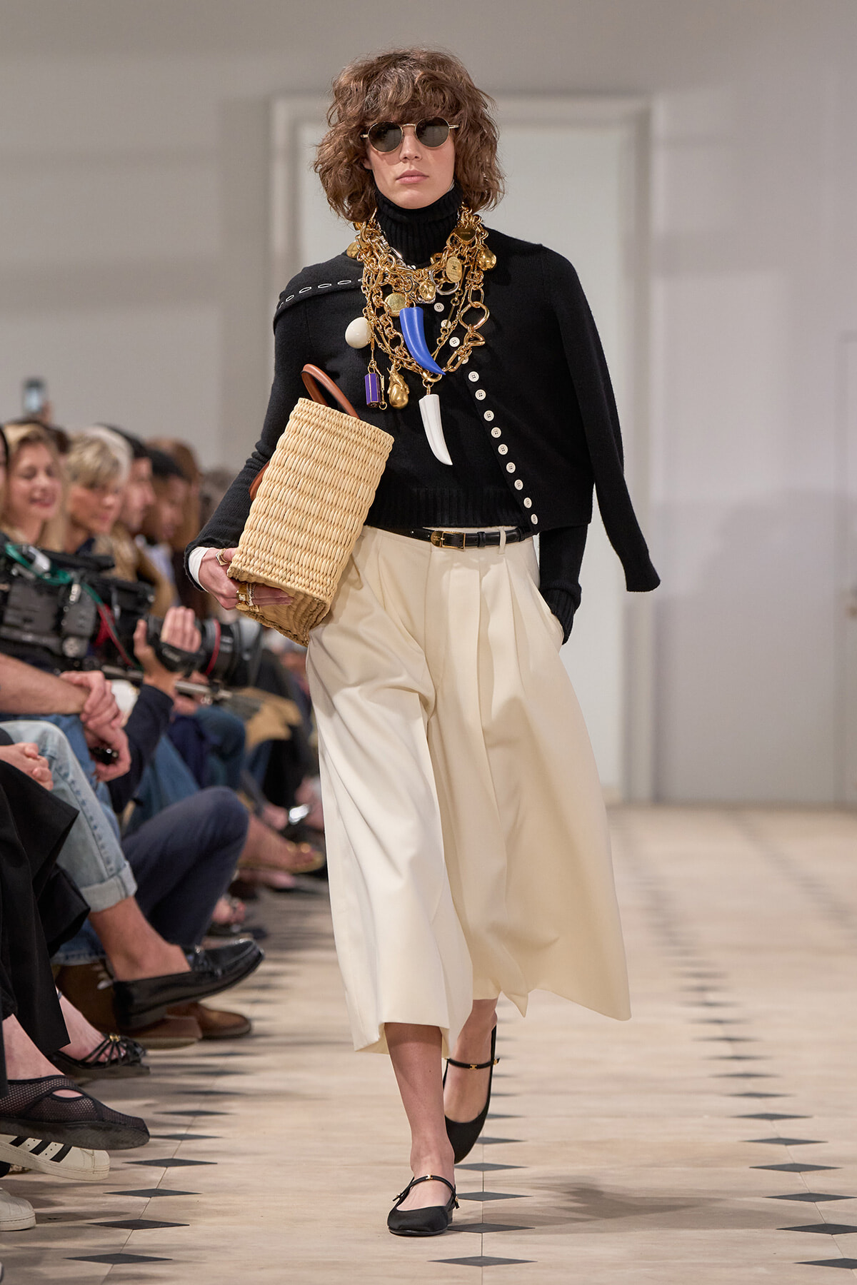 Model on runway wearing a black turtleneck sweater, cream wide-leg pants, oversized gold jewelry, sunglasses, and a woven straw handbag.