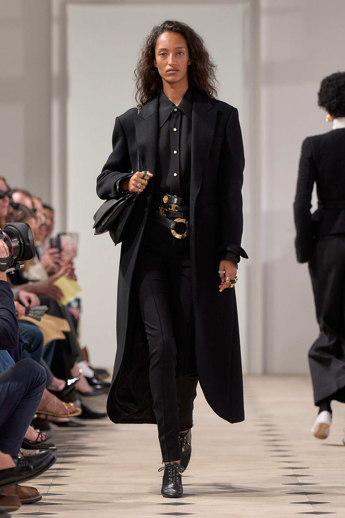 Model struts down a runway in an all-black ensemble: long black coat, black shirt, slim pants, and lace-up boots, accessorized with rings and belts. Audience watches.