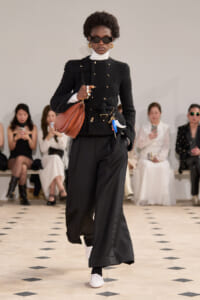 Model on a runway wearing a black boucle jacket with gold buttons, white turtleneck, wide‑leg black trousers, white sneakers, sunglasses, and a brown shoulder bag.