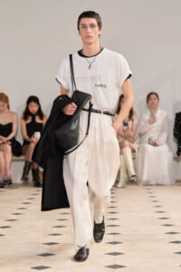 Male model on a runway wearing a white CELINE Paris tee tucked into white high-waisted pants, with a black crossbody bag and a black coat draped over his arm.