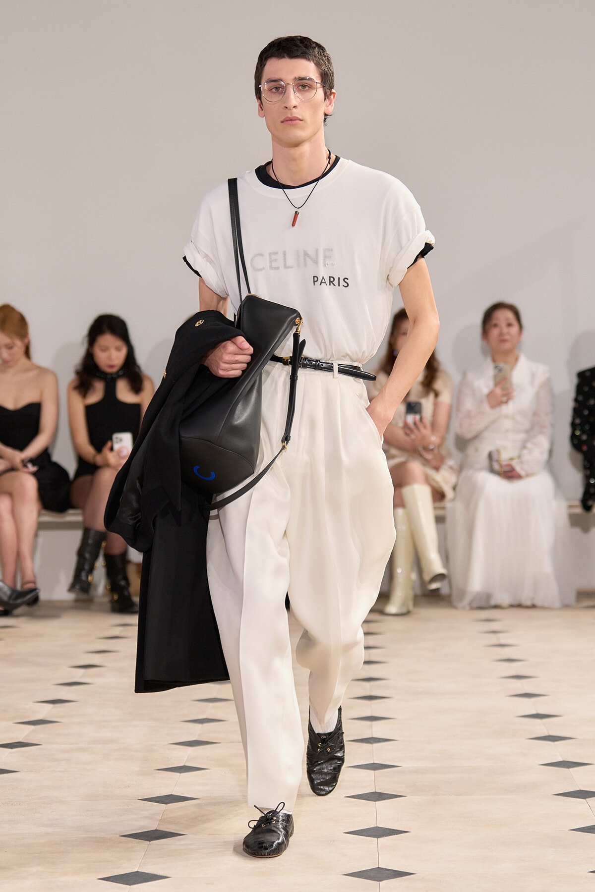 Male model on a runway wearing a white CELINE Paris tee tucked into white high-waisted pants, with a black crossbody bag and a black coat draped over his arm.