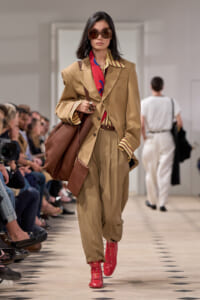 Model walking a runway in a tan suit with an oversized blazer, red scarf, large sunglasses, brown tote, and bright red mid-calf boots.