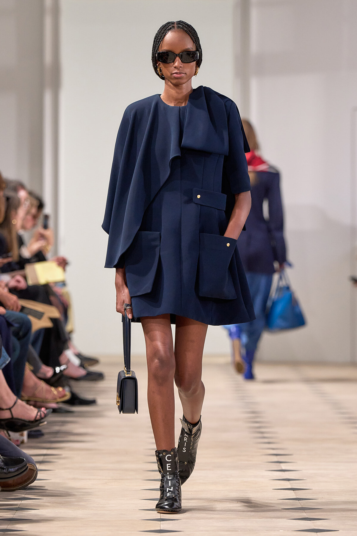 Model in a navy cape-style dress with oversized pockets walks the runway, carrying a small navy handbag and wearing black ankle boots with bold lettering.