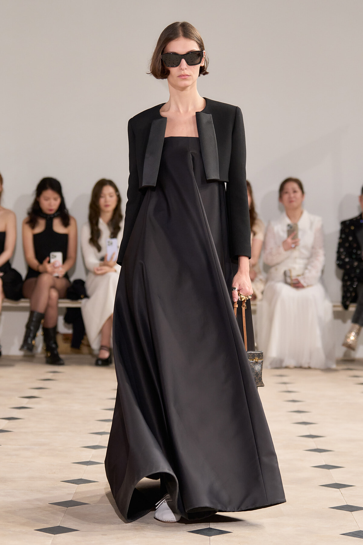 Runway model in a floor-length black strapless gown with a cropped black blazer, oversized sunglasses, and a small structured handbag.