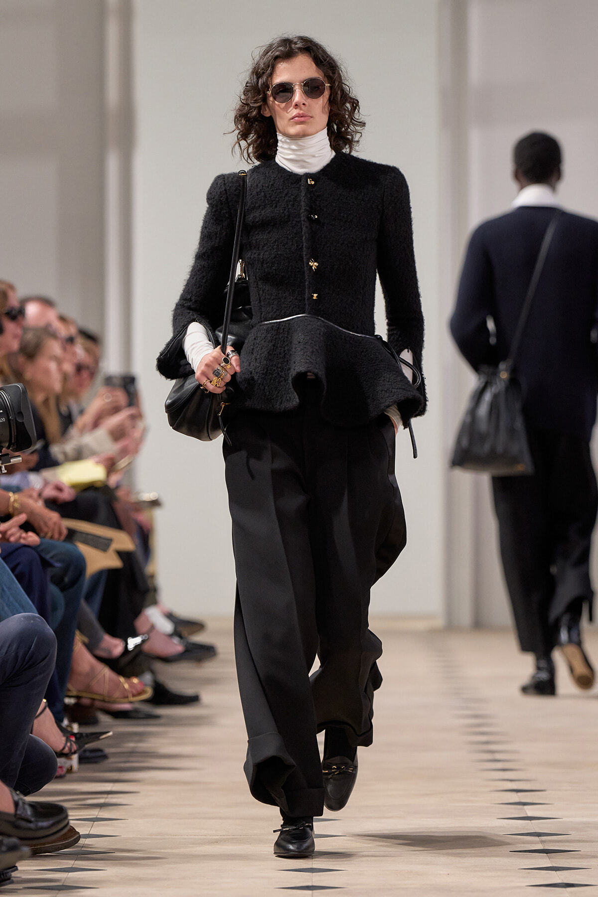 Model walking a fashion runway in a black textured peplum jacket, white turtleneck, black wide-leg pants, and sunglasses; audience seated on left.