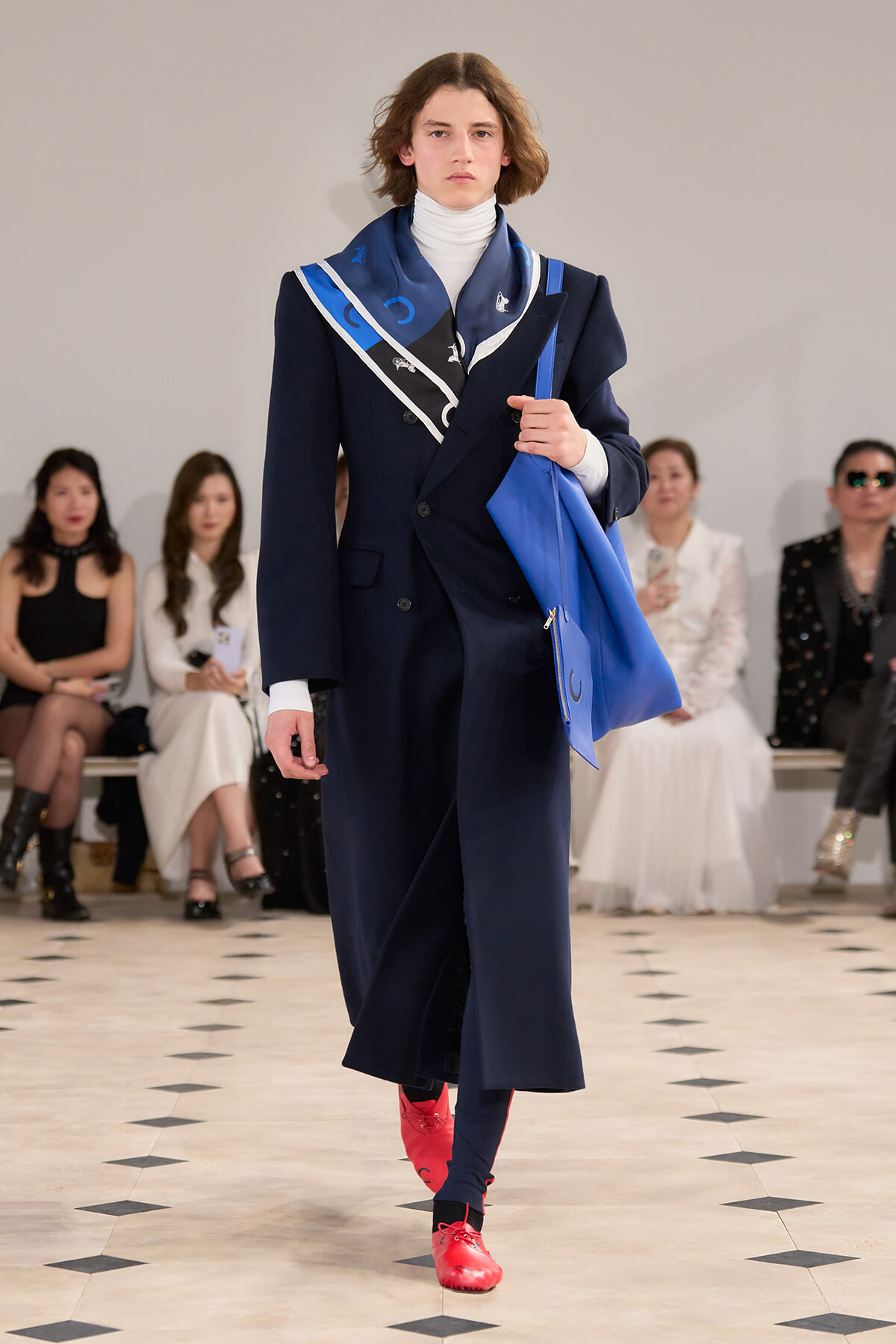 Model walking a fashion runway in a navy double-breasted coat, white turtleneck, and bright red shoes, carrying a blue tote bag