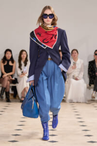 Runway model in a navy blazer, light blue pants, red scarf, oversized sunglasses, and cobalt CELINE boots, carrying a blue bag; audience in background.