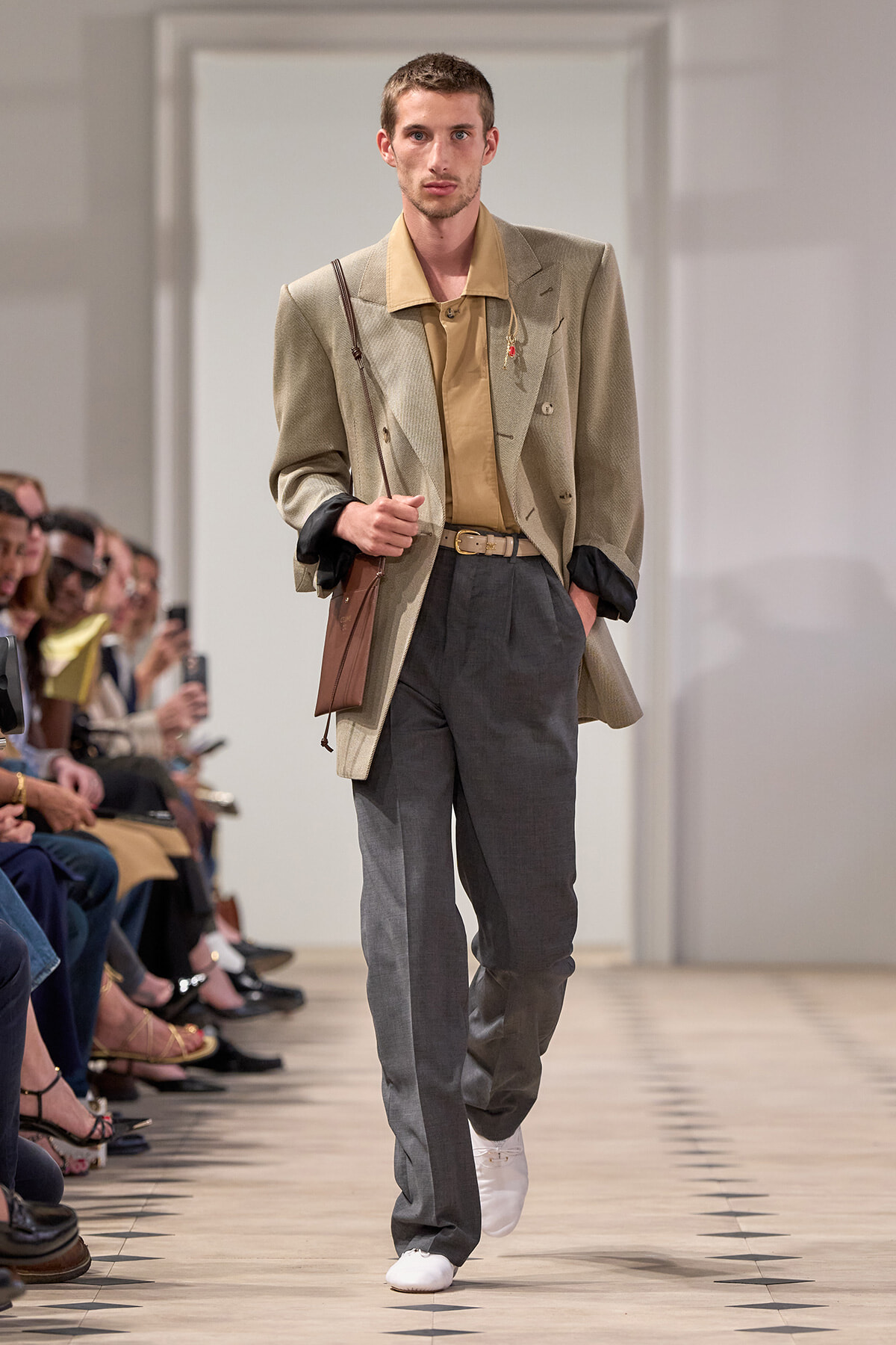 Male model walking a runway in a beige blazer, tan shirt, gray dress pants, white sneakers, and a brown crossbody bag; audience on the left.