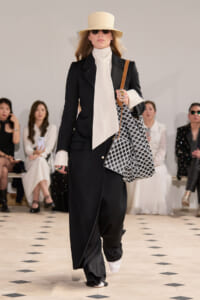 Runway model in a black blazer and wide-leg pants with a cream neck scarf, beige hat, sunglasses, and a black‑white patterned tote bag.