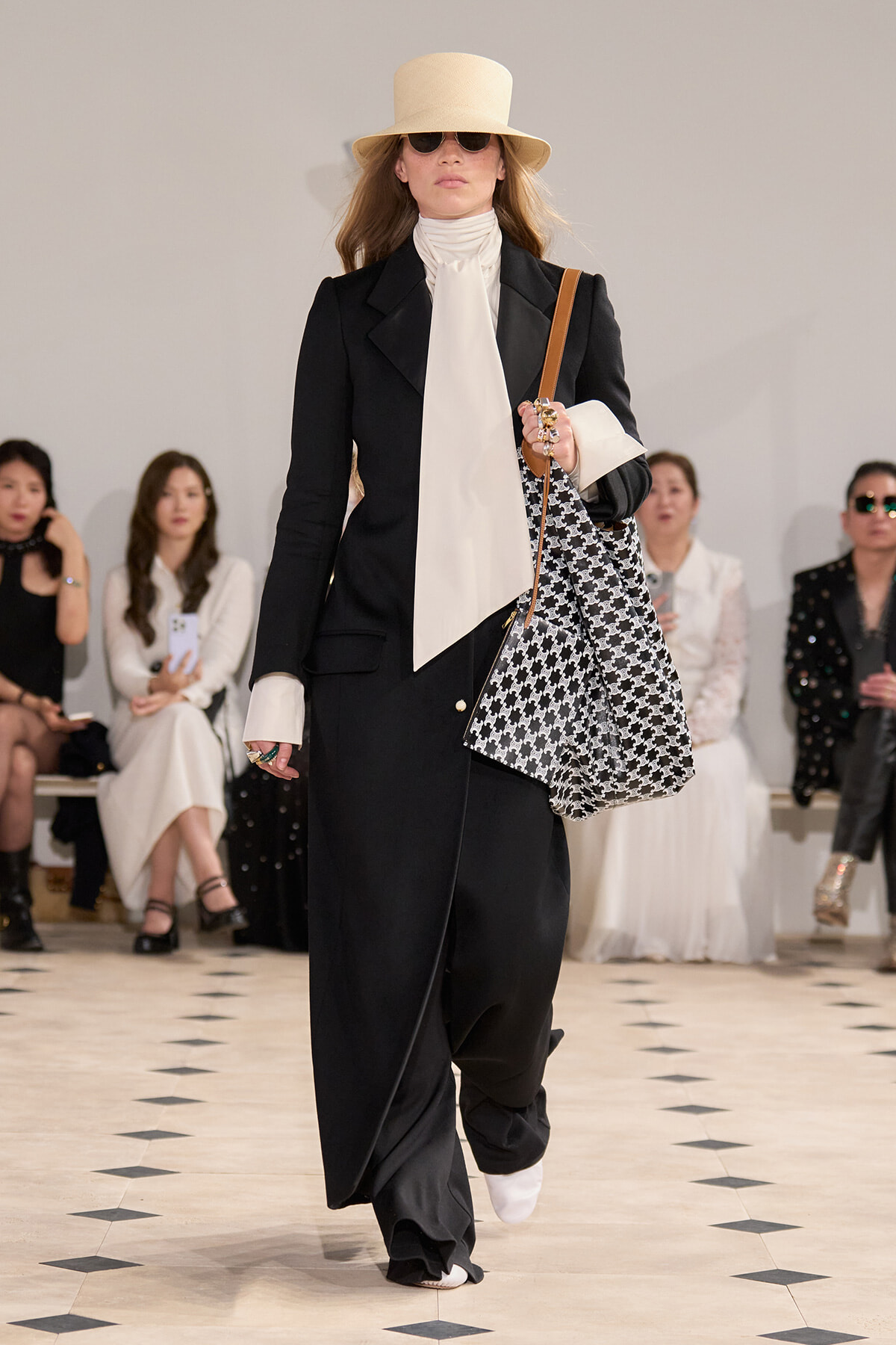Runway model in a black blazer and wide-leg pants with a cream neck scarf, beige hat, sunglasses, and a black‑white patterned tote bag.