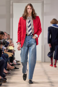 Model walking the runway in a red blazer, striped shirt, striped tie, and loose blue jeans with one hand in her pocket.