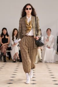 Model walking a fashion runway in a brown plaid suit with a tan scarf, white shirt, and white CELINE boots; carrying a black shoulder bag and wearing round sunglasses.