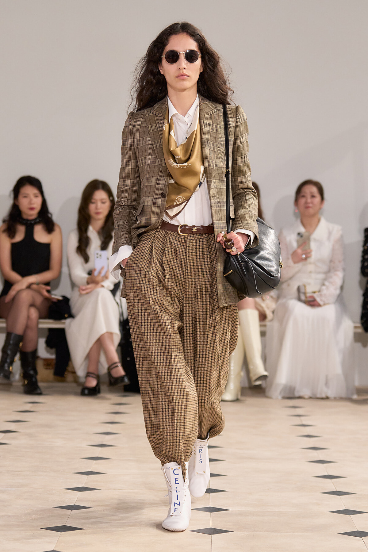 Model walking a fashion runway in a brown plaid suit with a tan scarf, white shirt, and white CELINE boots; carrying a black shoulder bag and wearing round sunglasses.