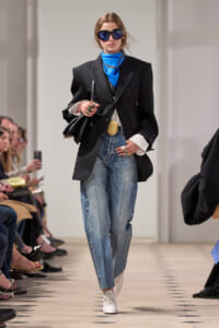 Model walks a fashion show wearing an oversized black blazer, bright blue neck scarf, high-waisted faded jeans, white lace-up shoes, dark sunglasses, and a black bag.