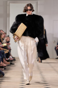 Model on a runway wearing a voluminous black fur top with white shirt, cream satin pants, beige loafers, and a woven straw clutch paired with dark sunglasses.