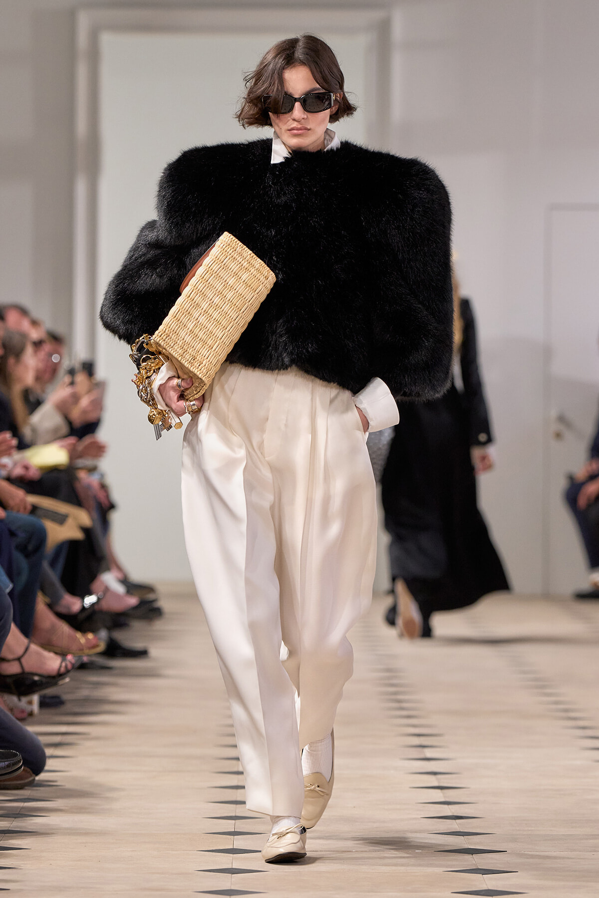 Model on a runway wearing a voluminous black fur top with white shirt, cream satin pants, beige loafers, and a woven straw clutch paired with dark sunglasses.