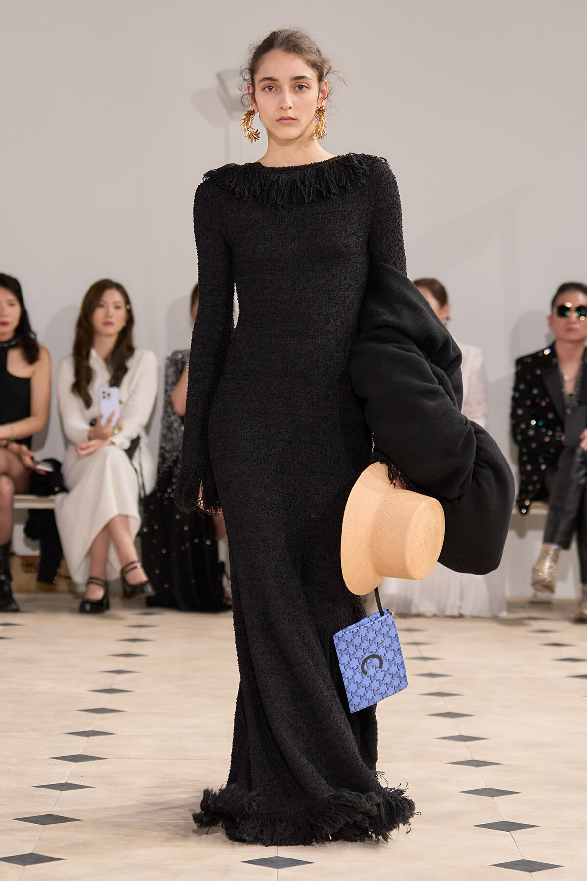 Model on a runway wearing a black textured fringe-hem dress, holding a beige hat and draping a black coat, with a blue patterned clutch visible.