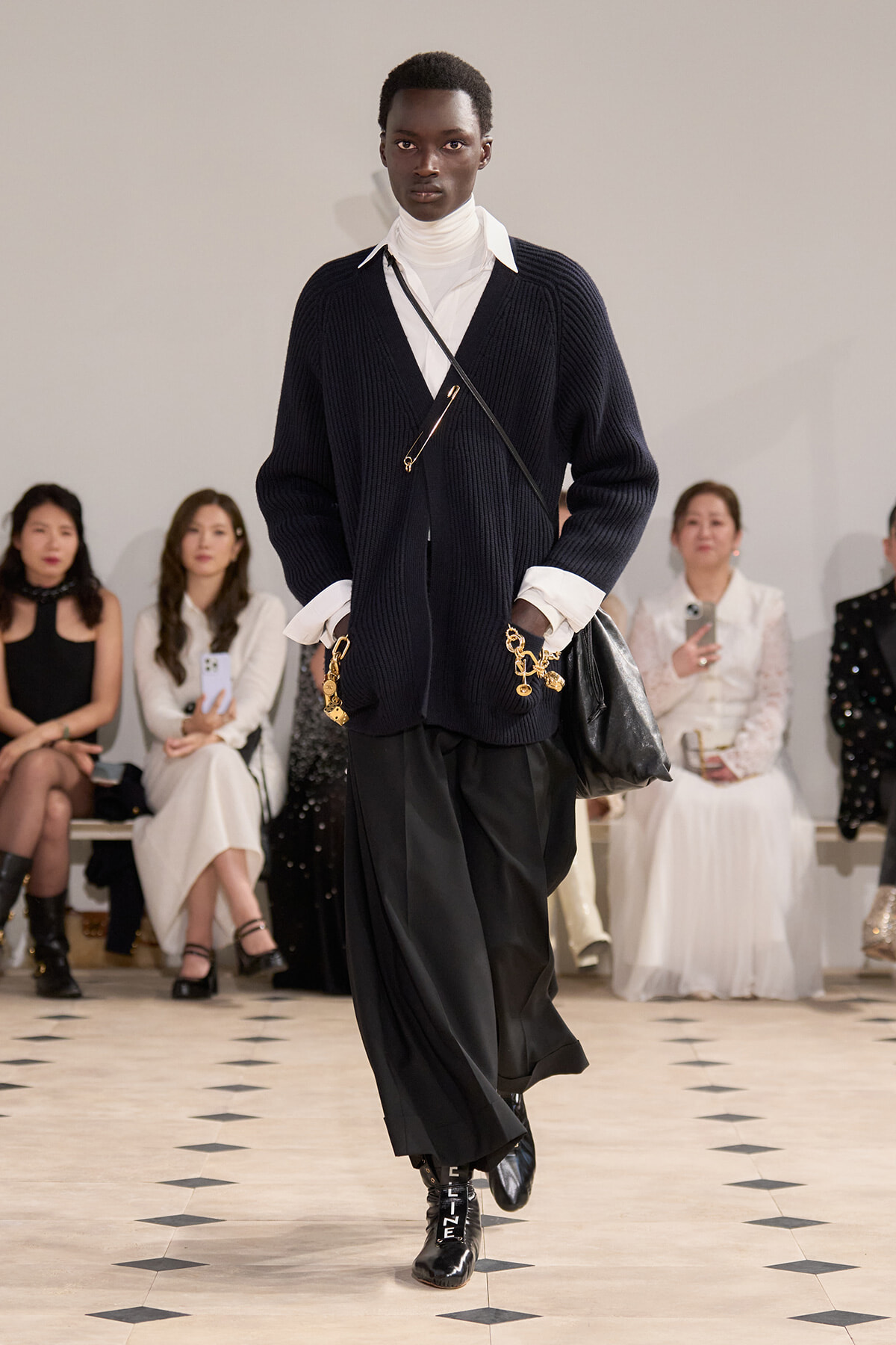 Model walking a runway in a navy cardigan over a white turtleneck, black wide-leg pants, and patent boots with gold accessories; audience seated in background