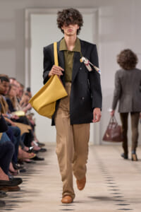 Male model on a fashion runway carrying a large yellow tote, wearing a black blazer, olive shirt, and tan trousers.