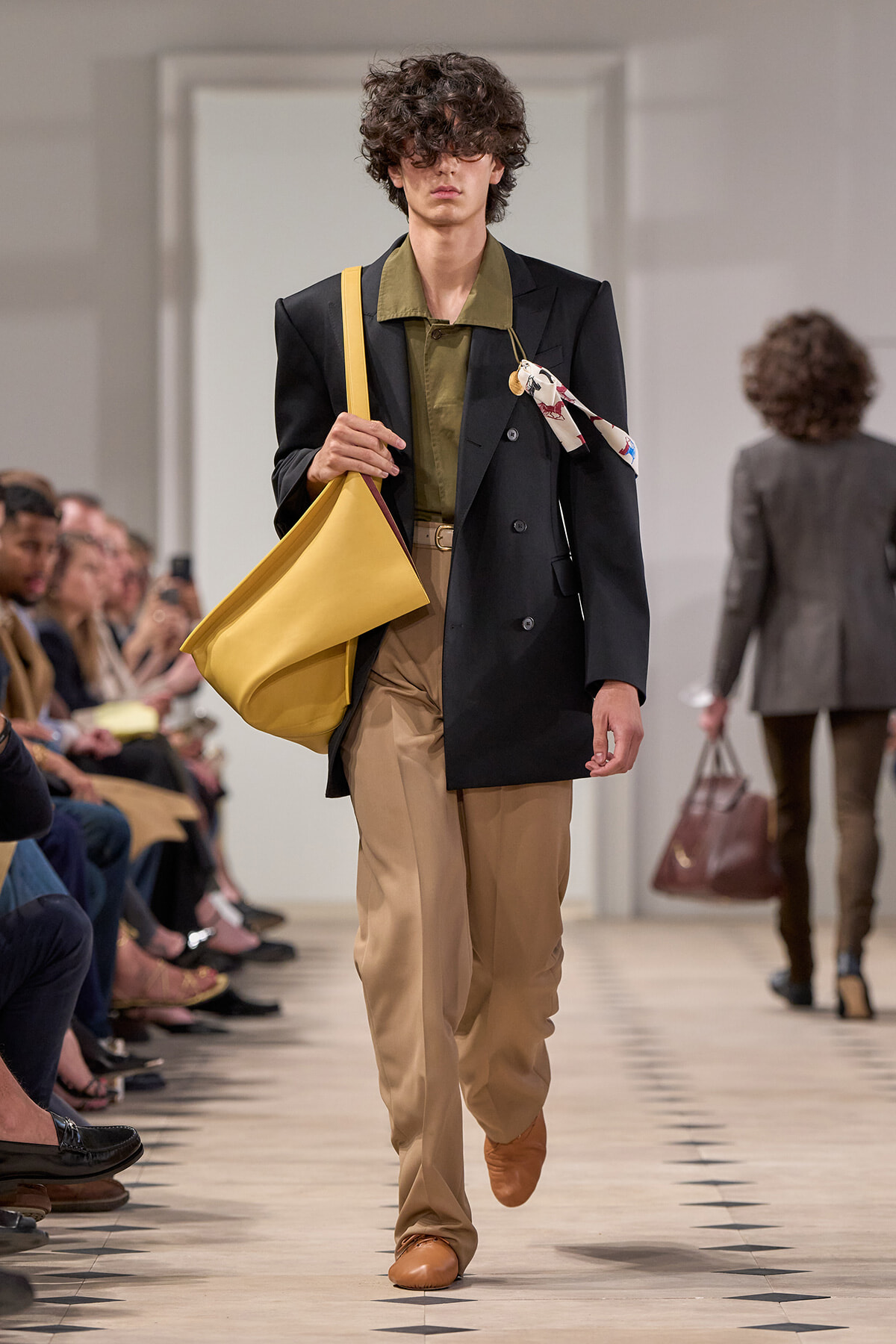 Male model on a fashion runway carrying a large yellow tote, wearing a black blazer, olive shirt, and tan trousers.