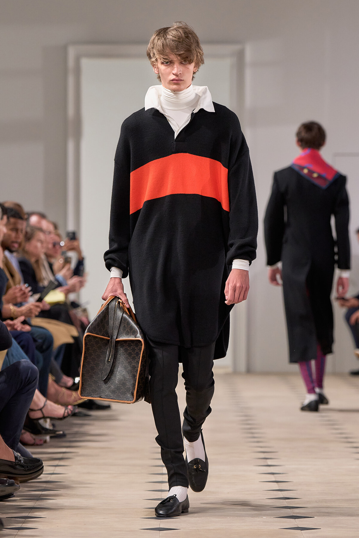 Male model on a fashion runway wearing a black sweater with a wide red stripe over a white turtleneck, carrying a large brown patterned bag.