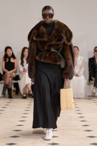 Model walks a runway in a brown fur coat over a black outfit, large sunglasses, black wide-leg pants, white sneakers, and a woven straw tote.