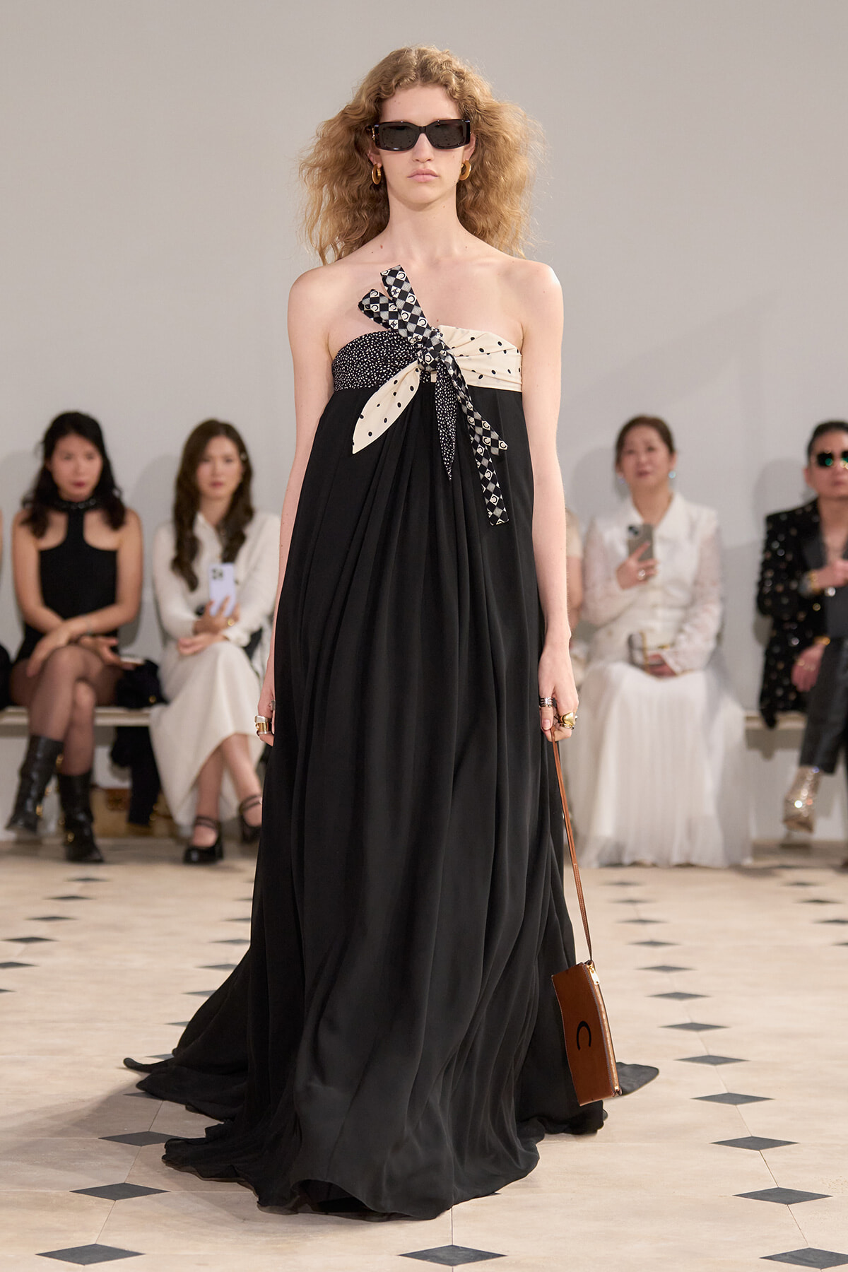 Model walks the runway in a black, one-shoulder gown with a patterned bow and beige band at the bodice.