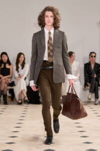 Male model walks the runway in a brown-toned outfit: tweed blazer, white shirt, patterned brown tie, slim brown pants, black shoes, and a large dark brown leather duffel bag.