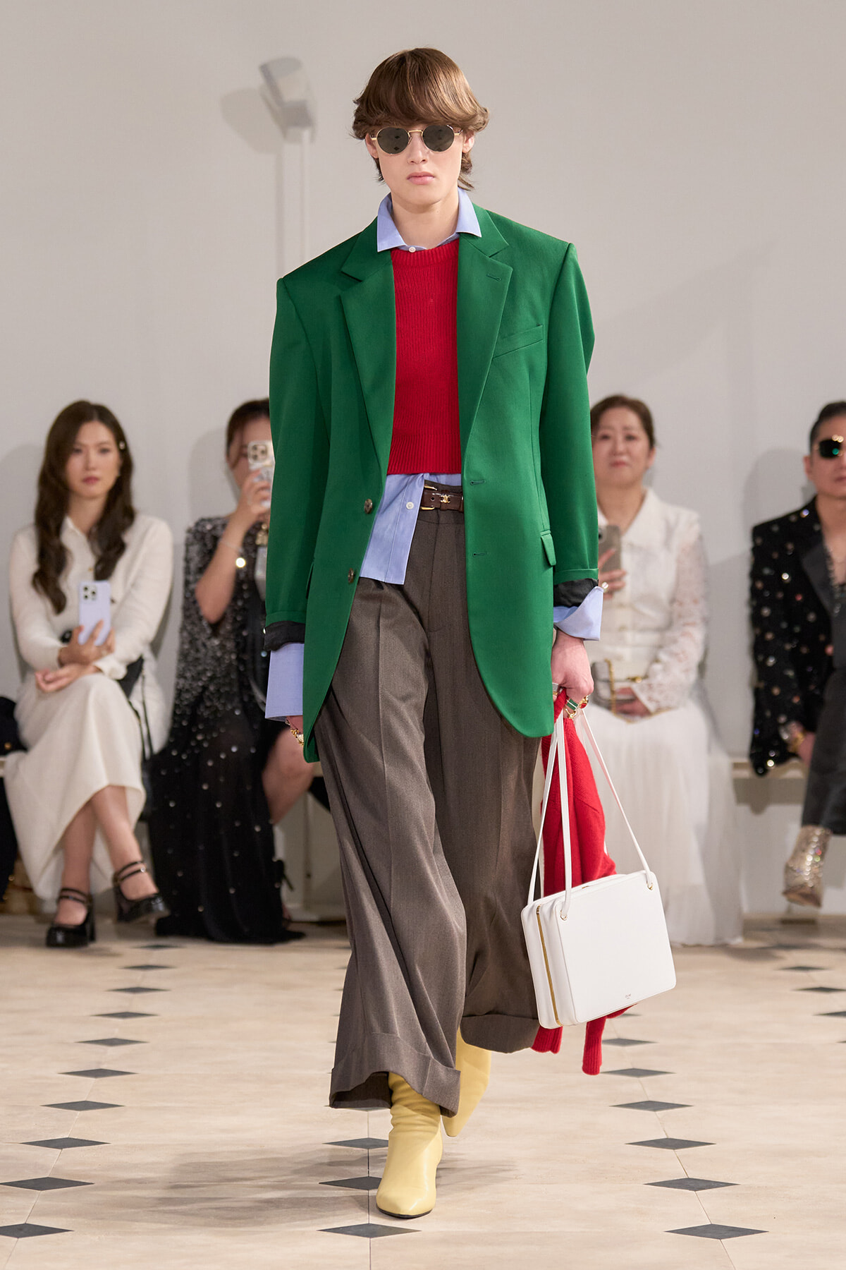 Model on a runway wearing a bright green oversized blazer over a red knit sweater and light blue shirt, gray wide-leg trousers, yellow ankle boots, round sunglasses, and carrying a white handbag.