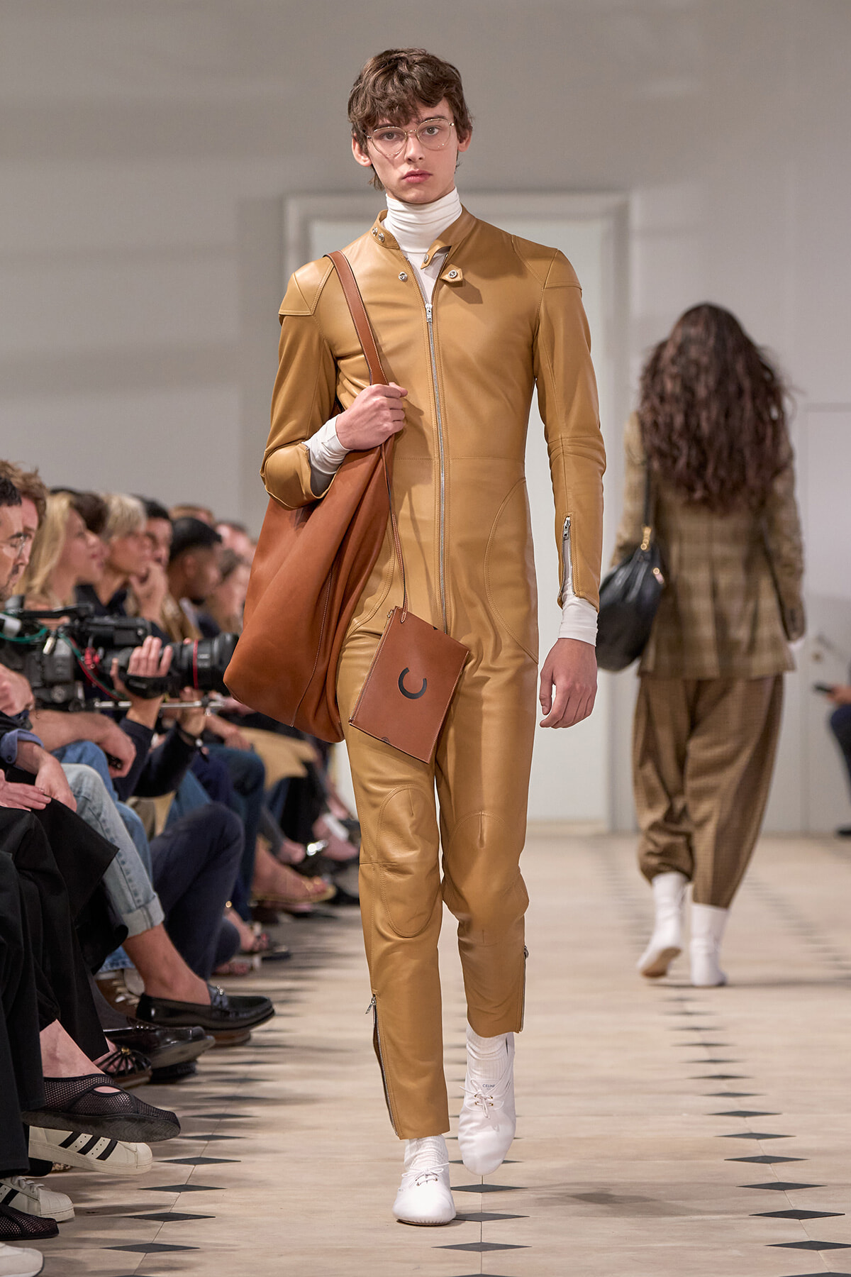Male model walking the runway in a tan leather jumpsuit with white turtleneck, carrying a brown slouch bag and a small square pouch, white sneakers, and clear-framed glasses, audience seated along the catwalk in background.