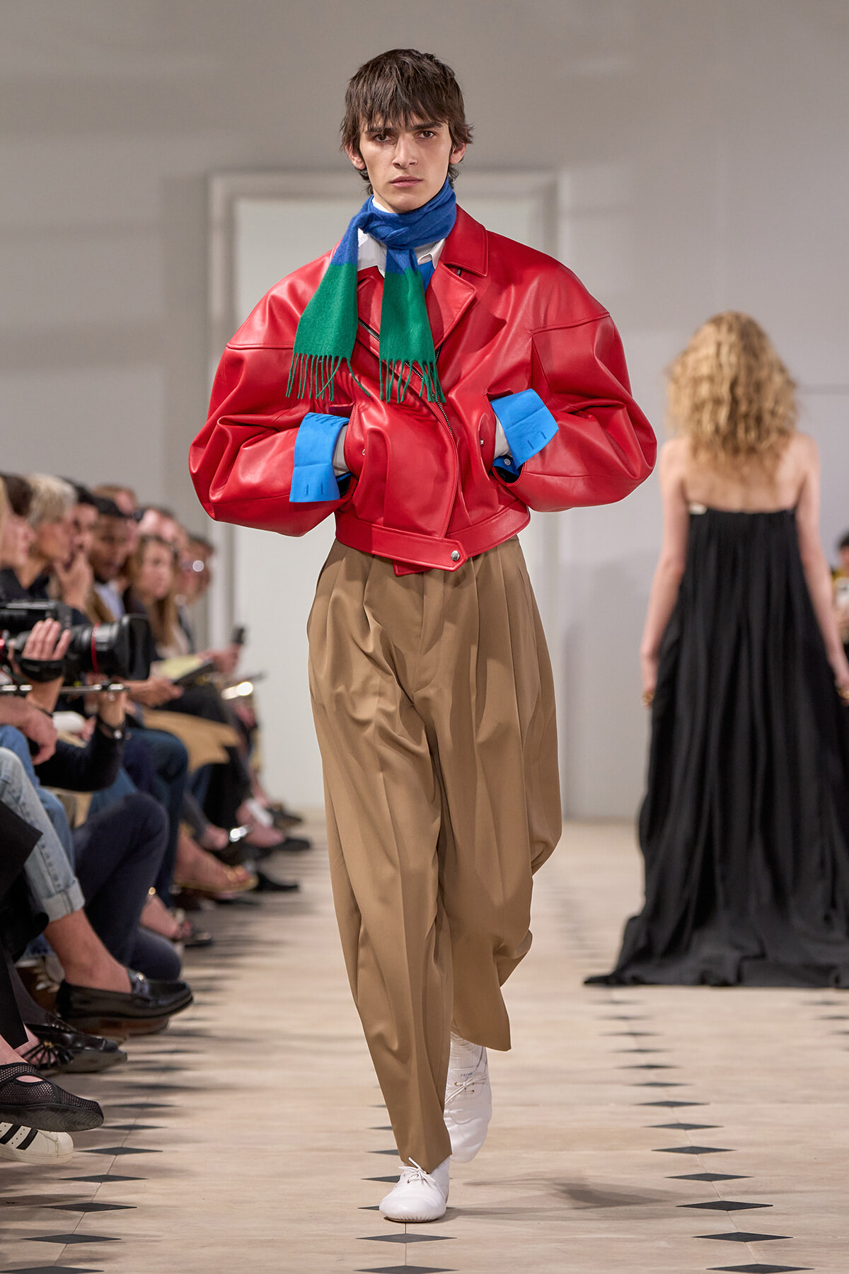 Runway model wearing an oversized red leather jacket, green and blue scarf, and tan wide-leg pants with white sneakers.
