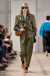 Model walks runway in olive suit with scarf, black sunglasses, and brown accessories; audience seated along the catwalk.