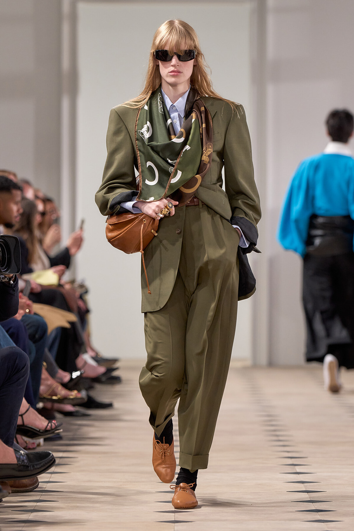 Model walks runway in olive suit with scarf, black sunglasses, and brown accessories; audience seated along the catwalk.