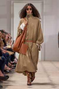 Model walking a runway in a tan belted trench cape, carrying a large brown tote and wearing patterned boots.