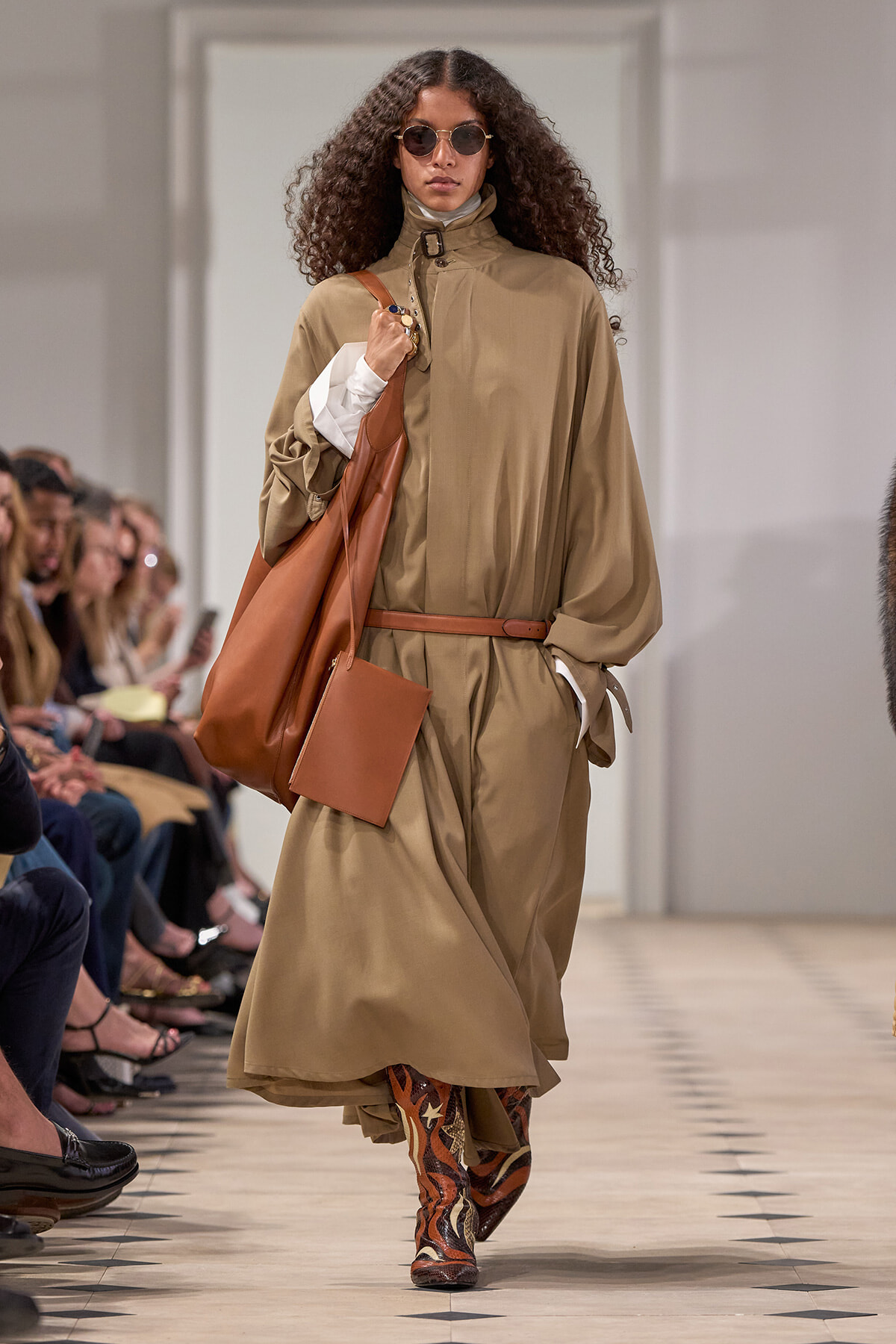 Model walking a runway in a tan belted trench cape, carrying a large brown tote and wearing patterned boots.