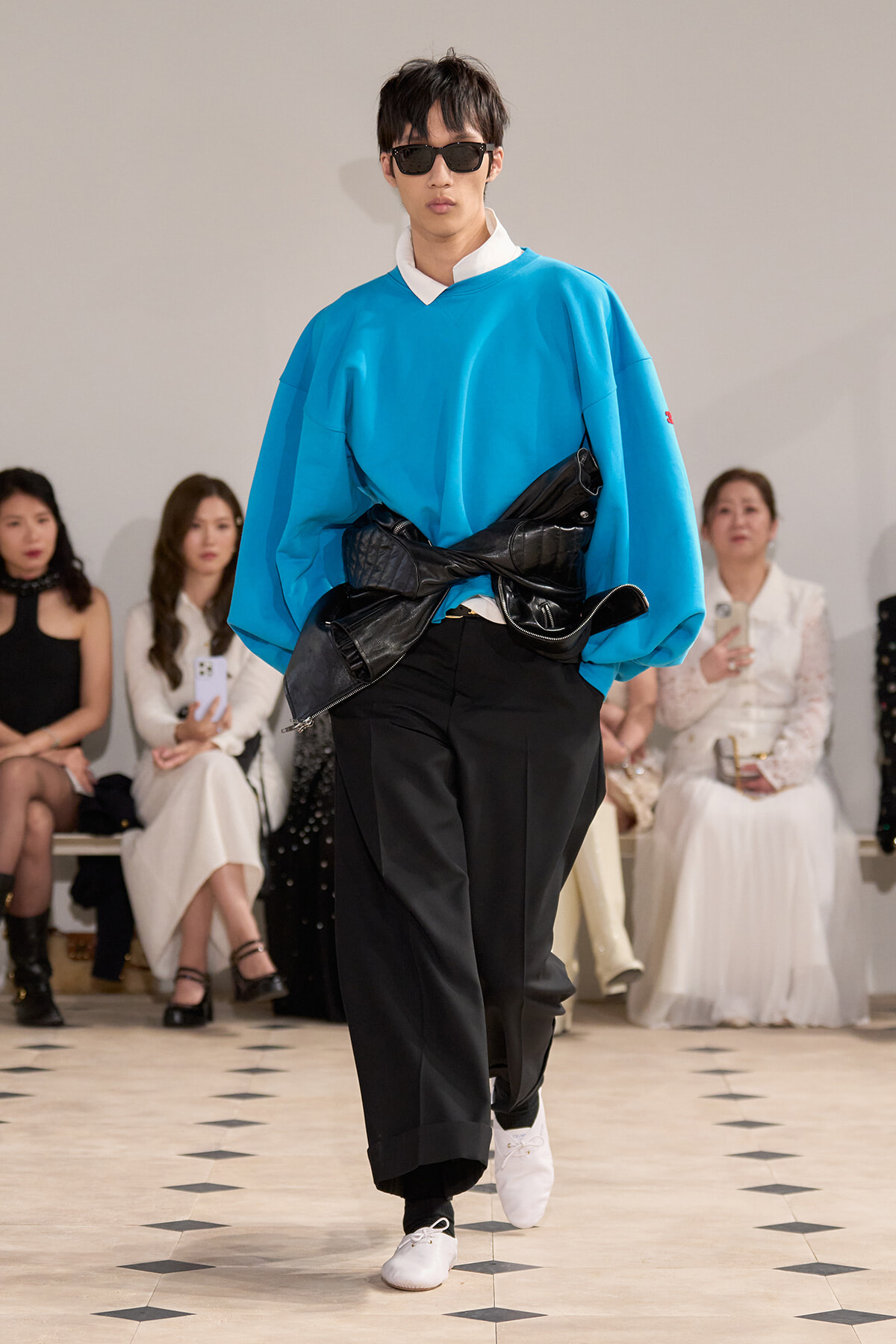 Male model walks the runway in a bright blue oversized sweatshirt with a white collar and a black wrapped beltbag over black wide trousers, wearing sunglasses.