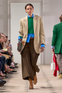Model walking a fashion show runway in a beige blazer, blue shirt, green knit sweater, and brown wide-leg trousers with tan boots.