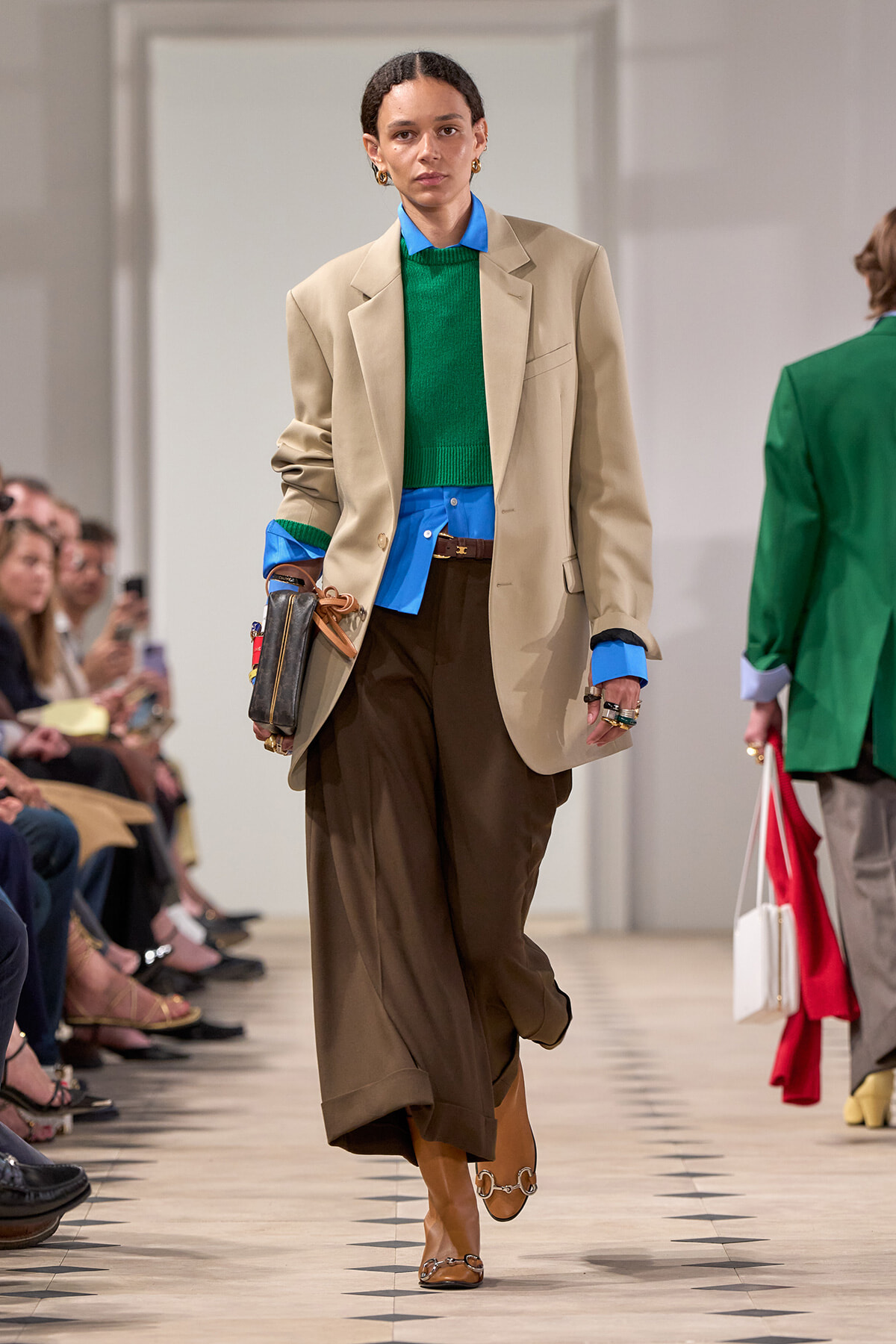 Model walking a fashion show runway in a beige blazer, blue shirt, green knit sweater, and brown wide-leg trousers with tan boots.