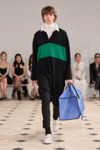 Male model walks a runway wearing a black sweater with a green stripe, white turtleneck, black pants, and white sneakers, carrying a large blue patterned bag.