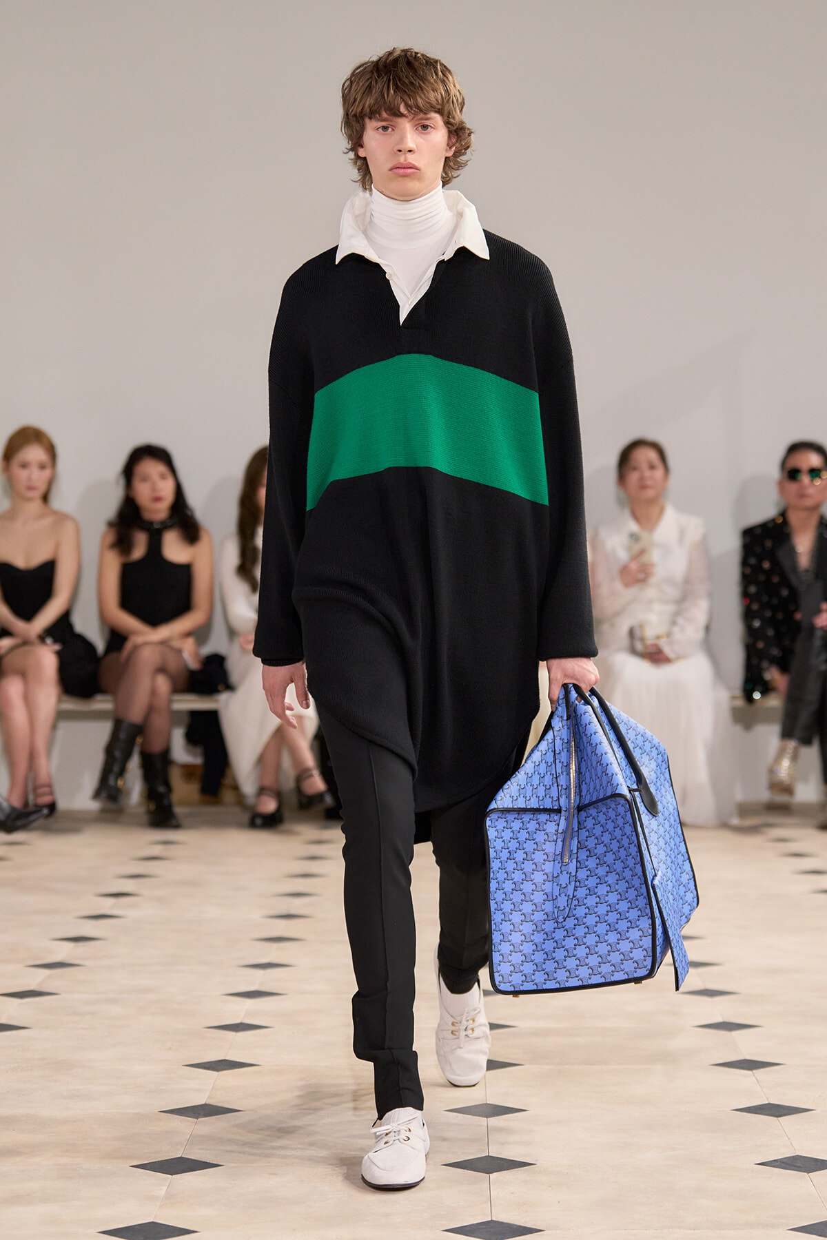 Male model walks a runway wearing a black sweater with a green stripe, white turtleneck, black pants, and white sneakers, carrying a large blue patterned bag.