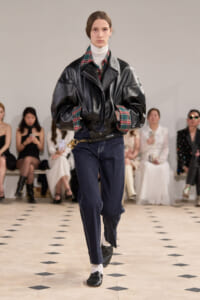 Model on a runway wearing an oversized black leather jacket over a plaid shirt and white turtleneck, with dark jeans and a CELINE belt.