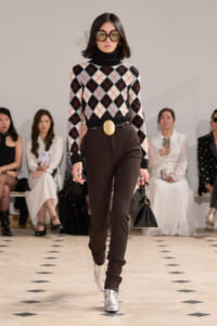 Model walking a fashion runway in a black turtleneck argyle sweater, brown tailored pants, oversized sunglasses, a gold belt buckle, and silver boots.
